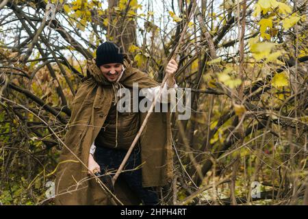 Porträt eines lächelnden Touristen-Mannes im Regenmantel-Zelt, der kleine Äste mit einer kleinen Schaufel im Wald an bewölktem, kaltem Tag hackt. Stockfoto