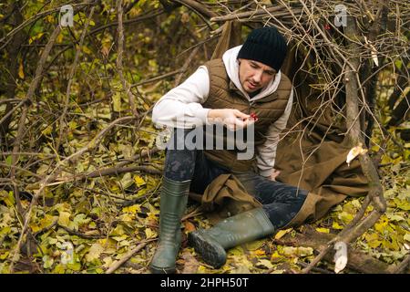 Hungerwanderer, der im Dickicht unter einem Baum im Wald auf einem Regenmantel sitzt und wilde rote Beeren in den Händen hält und sie an bewölktem regnerischen Tag ansieht. Stockfoto