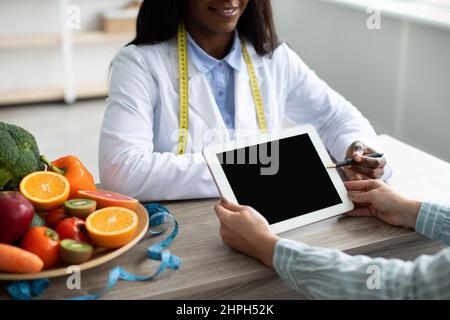 Schwarze Ernährungsberin zeigt auf eine Tablette mit leerem Bildschirm, erklärt den Diätplan und gibt Ratschläge für die Patientin Stockfoto