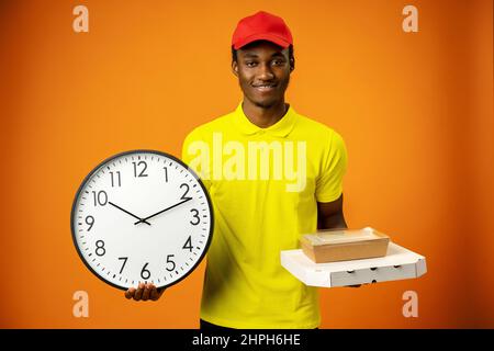 Lächelnder afroamerikanischer Kurier mit Uhr und Box auf orangefarbenem Hintergrund Stockfoto
