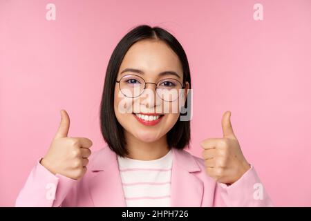 Super, Glückwunsch. Gesicht einer aufgeregten asiatischen Geschäftsfrau in einer Brille, lächelnd zufrieden, mit Daumen nach oben, stehend über rosa Hintergrund Stockfoto