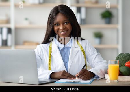 Freundlicher afroamerikanischer Ernährungsberater, der einen Speiseplan für den Patienten erstellt, im medizinischen Büro arbeitet und vor der Kamera lächelt Stockfoto