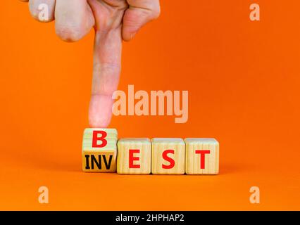 Symbol für beste Investition. Geschäftsmann dreht einen Holzwürfel und ändert das Wort Invest in Best. Schöner orangefarbener Tisch, orangefarbener Hintergrund. Business und Best Stockfoto