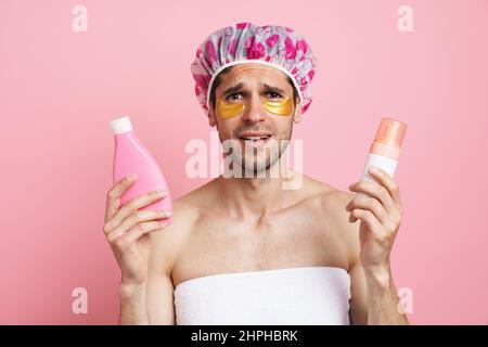 Junger weißer Mann mit Duschhaube posiert mit Beauty-Produkten isoliert auf rosa Hintergrund Stockfoto