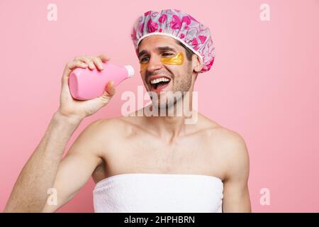 Junger weißer Mann mit Duschhaube, Shampoo haltend und isoliertem Gesang auf rosafarbenem Hintergrund Stockfoto