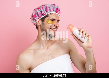 Junger weißer Mann mit Duschhaube, Shampoo haltend und isoliertem Gesang auf rosafarbenem Hintergrund Stockfoto