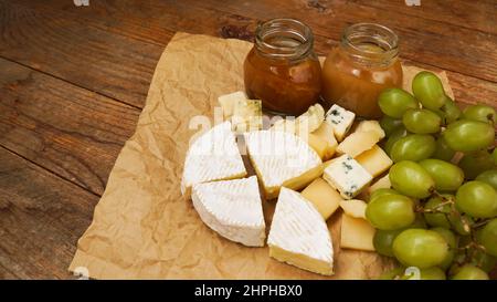 Camembert und andere Arten von Käse und grünen Trauben auf Bastelpapier mit hausgemachter Marmelade und Honig im Glas, Lebensmittelkonzept Stockfoto