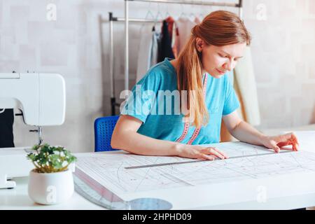 Ein weiblicher Schneider macht ein Muster. Junge lächelnde Modedesignerin, die über dem Schreibtisch an einer Nähmaschine arbeitet. Schneiderin, die in ihrer Werkstatt ein Kleidungsstück herstellt Stockfoto