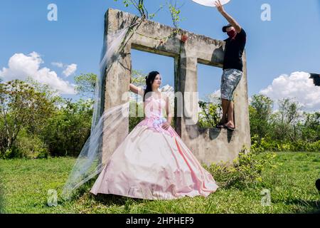 Das 15-jährige kubanische Mädchen posiert mittags für ein Foto im Freien und trägt ein langweitiges, altmodisches Kleid. Es gibt einen jungen Mann, der einen hausgemachten Diff hält Stockfoto