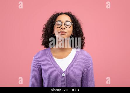 Ruhige junge schwarze Frau in einer Brille, die ihre Augen schließt und auf pinkem Studiohintergrund frei atmet Stockfoto