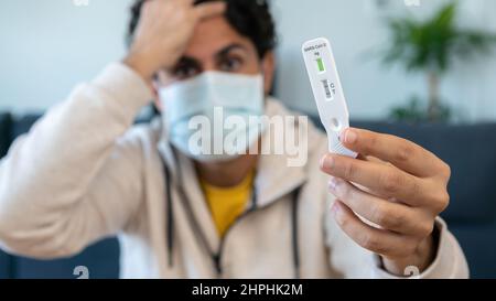 Ein trauriger Kaukasusmann stellt ein Schnelltestgerät für das Covid19-Virus her, das ein positives Ergebnis zeigt und seinen Kopf an einem Holztisch hält. Antigenkasset Stockfoto