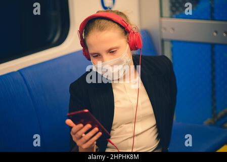 Nettes, ziemlich besorgtes junges Mädchen mit Maske und Kopfhörern in Bus, Zug oder U-Bahn, das zur Schule geht Stockfoto