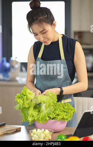 Porträt einer schönen asiatischen Frau mit Schürze, die ihr grünes Gemüse für ihre gesunde Salatschüssel in der Küche zubereitet. Stockfoto