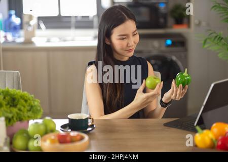 Charmante asiatische Frau auf Tablet-Bildschirm und hält einen grünen Apfel und einen grünen Pfeffer in der Küche, Online-Kochen lernen, Kochen Blogger. Stockfoto