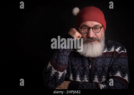 Reifer bärtiger Kaukasusmann sieht in der Kamera mit einer Brille auf dem Gesicht aus, Mann mit rotem weihnachtsmann-Hut, schwarzem Hintergrund Stockfoto