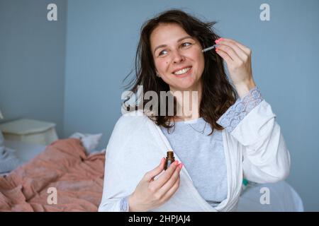 Frau, die mit einer Anti-Absackpipette und Serum-Essence-Öl eine saubere Augenkontur berührt und im Schlafzimmer auf dem Bett im Bademantel wegschaut. Konzept Faltenpflegeprodukte in Faltenbildung zu Hause Stockfoto