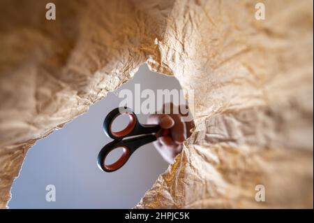 Eine Hand legt eine Schere in einen Papierbeutel. Die Hand eines reifen Mannes hält eine Büroschere über einer offenen braunen Tasche. Von unten nach oben geschossen. Nahaufnahme. Selektiv Stockfoto