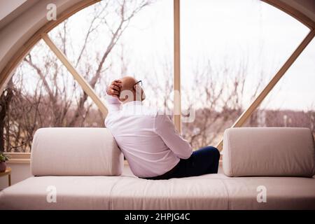 Rückansicht eines erschöpften Mannes, der auf dem Sofa saß und aus dem Fenster schaute. Stockfoto