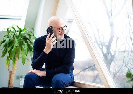 Aufnahme eines gutaussehenden Mannes mit Rollkragen-Pullover und Brillen, der am Fenster saß und einen Anruf entgegennehmen musste. Stockfoto