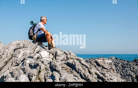 Älterer Mann, der auf Felsen sitzt und die Meereslandschaft betrachtet Stockfoto