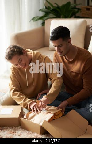 Glückliche junge Frau Auspacken der Box mit Kleidung mit Mann sitzt in der Nähe von ihr und helfen, sie bekommen Lieferung nach Hause Stockfoto