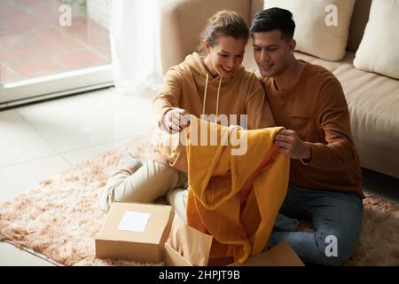 Das junge Paar bewunderte den neuen Kapuzenpullover in ihren Händen, während sie auf dem Boden im Zimmer saßen und ihr Paket nahmen Stockfoto