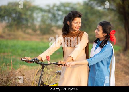 Glückliche junge indische Mädchen in Schuluniform stehen mit dem Fahrrad auf der Dorfstraße und schauen sich an und lächeln. Stockfoto