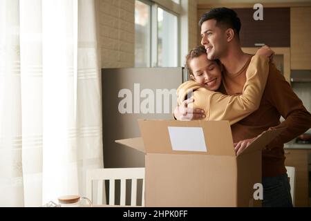 Glückliche junge Frau umarmt ihren Freund, während sie zu Hause in der Küche stehen und das große Paket erhalten Stockfoto