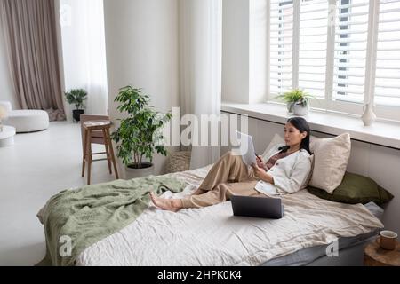 Junge, selbstbewusste asiatische Buchhalterin in legerer Kleidung, die auf dem Bett sitzt und Finanzpapiere durchschaut, während sie von zu Hause aus arbeitet Stockfoto