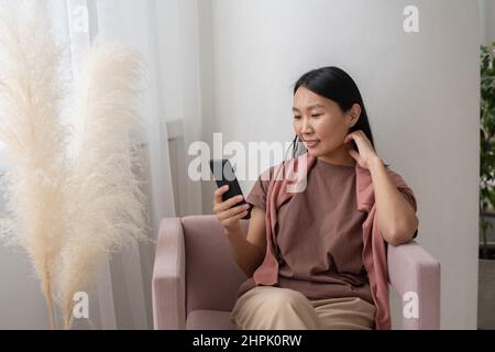 Junge lächelnde Asiatin in Casualwear sitzt im Sessel und schreibt im Smartphone oder schaut sich im Online-Shop Waren an Stockfoto