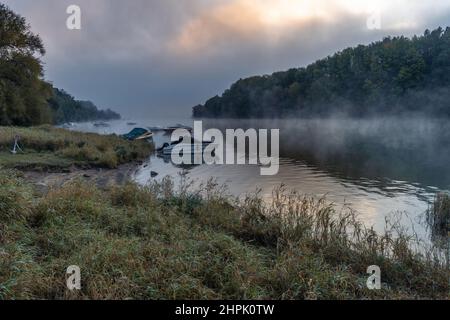 Ufer eines Sees am frühen Morgen des Herbstes. Der Nebel lichtet sich und erlaubt Einblicke in festfahrende Boote. Die erste Sonne des Tages erhellt die tiefen Wolken mit warmen Farbtönen, die sich im Wasser spiegeln. Stockfoto