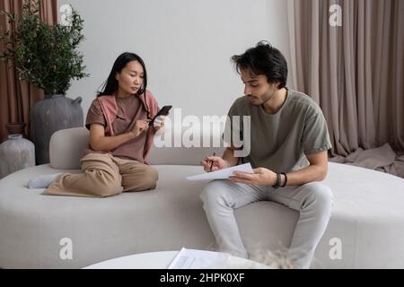 Junge Geschäftsmänner in legerer Kleidung analysieren Finanzdaten, während asiatische Frauen im Heimbüro auf dem Sofa SMS schreiben Stockfoto