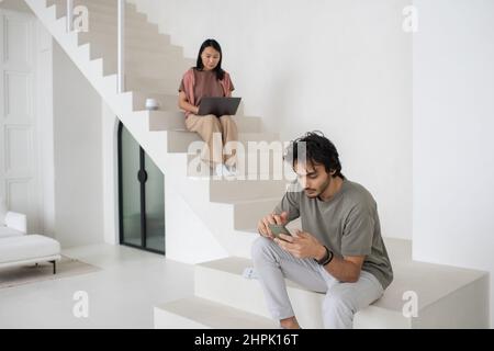 Junger kaukasischer Mann mit Smartphone sitzt auf einem weißen Koffer gegen asiatische Frauen mit Laptop, während beide von zu Hause aus arbeiten Stockfoto