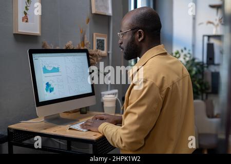 Ein moderner afroamerikanischer Geschäftsmann sitzt am Schreibtisch vor einem Computermonitor und schaut sich das Finanzdiagramm auf dem Bildschirm an Stockfoto