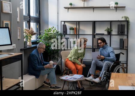 Zwei junge, multirassische Mitarbeiter besprechen das Dokument bei einem Arbeitstreffen im Büro vor einem reifen männlichen Kollegen mit Laptop Stockfoto