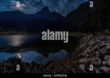 Nachtszene in den Schweizer Alpen. Das Wasser eines Bergsees reflektiert den Mond, der sich mit den Wolken am Himmel vermischt. Sie können die Silhouette des Berges und der Bäume sehen, die den See umgeben. Stockfoto