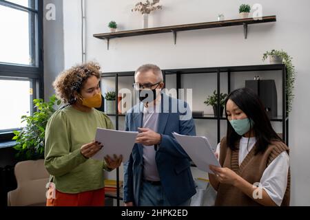 Reifer Geschäftsmann in Maske zeigt auf Dokument während der Diskussion mit jungen biracial weiblichen Kollegen bei der Arbeitssitzung im Büro Stockfoto