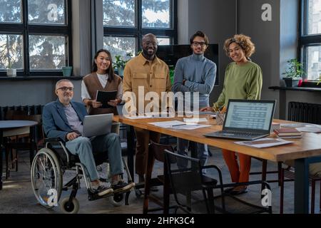 Fröhliche Ökonomen verschiedener Ethnien versammelten sich am Arbeitsplatz und schauten lächelnd auf die Kamera, während sie im Büro eine Arbeitssitzung hatten Stockfoto