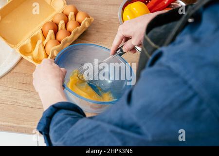 Draufsicht auf einen nicht erkennbaren Mann im blauen Langarm-Hemd, der Eier in einer blauen Kunststoffschüssel mit Drahtbesen auf dem Tisch in der Nähe eines Stahlsieders mit rotem, brüllenden Schrei schlägt Stockfoto
