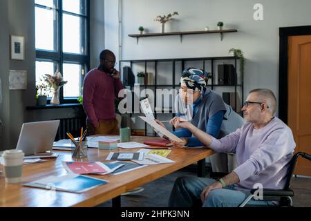 Reifer Geschäftsmann im Rollstuhl, der auf ein Dokument verweist, das von einer jungen biracial Kollegin während der Diskussion über Finanzdaten gehalten wurde Stockfoto