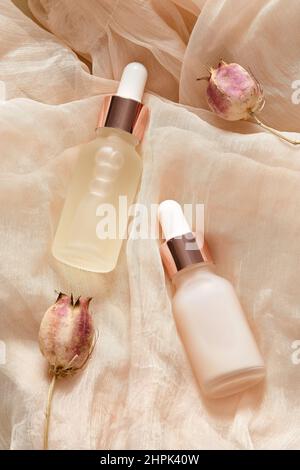 Transparente Tropfflaschen mit ätherischem Öl und Serum, getrocknete Blüten auf beigem Tuch. Aromaölflaschen Draufsicht. Stockfoto