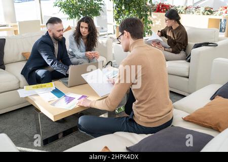 Junger Manager eines modernen Unternehmens, der Daten in Dokumenten analysiert, während er vor zwei Kollegen am Tisch sitzt, die über ein neues Projekt sprechen Stockfoto