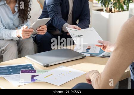 Ein Geschäftsmann, der während eines Arbeitsmeetens im Büro einen Finanzbericht an einen Kollegen über den Tisch mit Dokumenten und Laptop übergibt Stockfoto