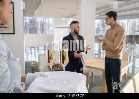 Junge Kellnerin in weißen Handschuhen mit Tablett und zwei Sektflöten für Mitarbeiter, die vor einer geschäftlichen Veranstaltung über ihre Arbeitspunkte sprechen Stockfoto