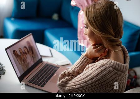 Porträt einer jungen blonden lächelnden Frau, die Fotos von Freunden vom Geburtstag auf dem Laptop zu Hause ansieht. Remote Festival Stockfoto