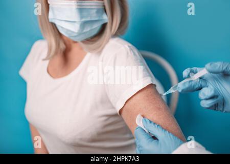 Impfung älterer Menschen. Der Arzt macht eine Impfung gegen ältere Frauen, die Gesichtsmaske auf blauem Hintergrund trägt. Stockfoto