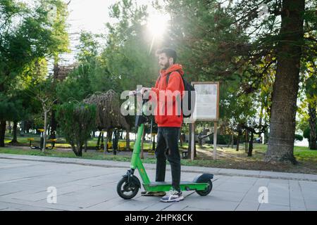 Elektroroller, junger Mann, der mit dem Roller im Park im Hintergrund steht. Spaß beim Fahren Elektroroller durch die Stadt. Stockfoto