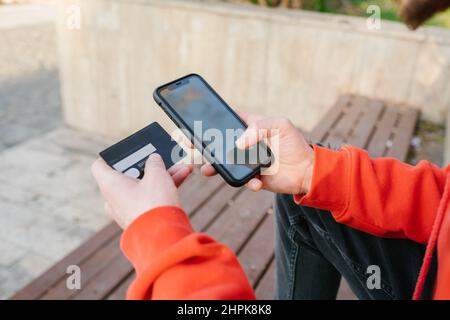 Mann mit Kreditkarte und Smartphone. Online etwas von außen kaufen. Der junge Mann sitzt in einem Park und kauft ein. Stockfoto