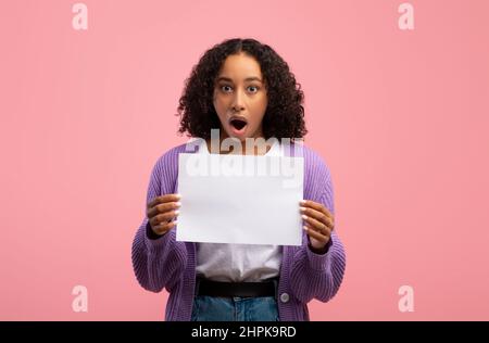 Emotionale junge schwarze Frau hält ein leeres Papierbanner, öffnet den Mund in Schock auf rosa Studiohintergrund, Nachbildung Stockfoto