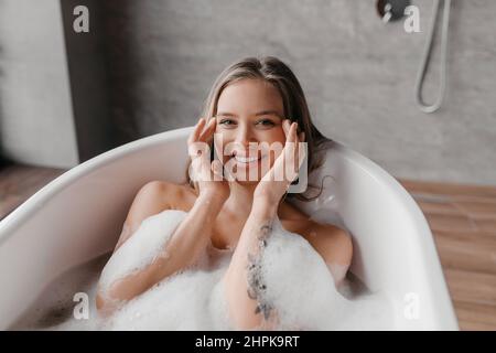 Hautpflege und Spa-Behandlungen zu Hause. Glückliche, entspannte Dame in der Badewanne mit Schaumstoff, berührte ihre glatte Gesichtshaut und lächelte Stockfoto